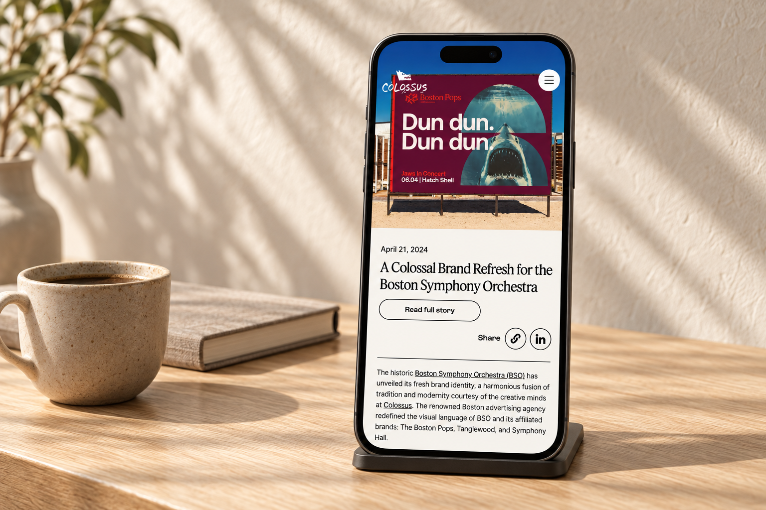 Colossus news article on a smartphone in a warm, minimal oak and cream workspace